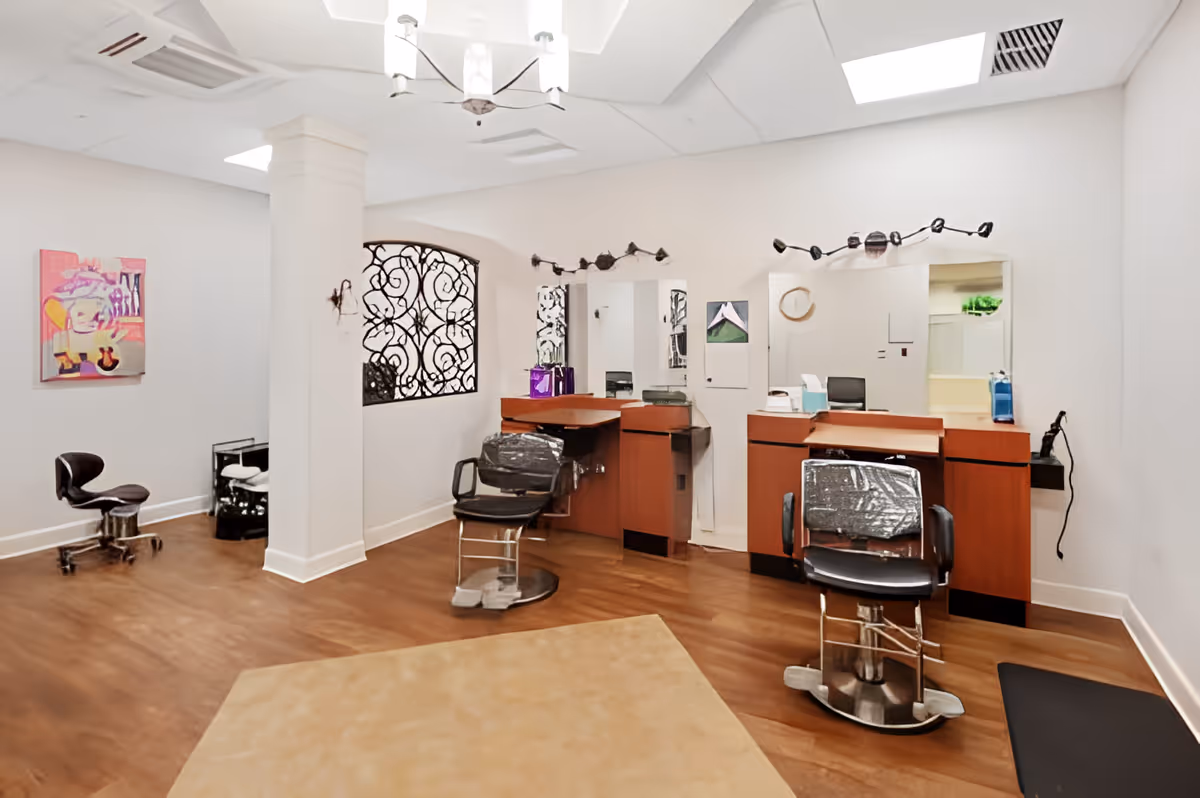 Interior view of a salon area with two black salon chairs in front of wooden counters with mirrors. The room has white walls, a decorative wrought iron window, a chandelier, and a piece of colorful artwork on the wall. The floor is wooden with a beige rug in the center.