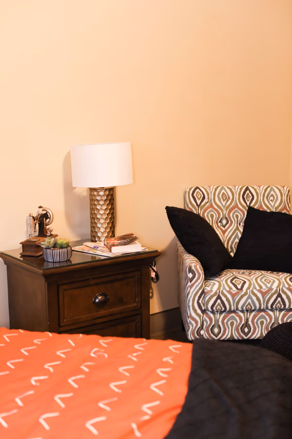 A cozy corner of a bedroom featuring a wooden nightstand with a decorative lamp, a small vintage sewing machine, a succulent plant, and some magazines. Next to the nightstand is a patterned armchair with two black cushions. The bed with an orange and black patterned blanket is partially visible in the foreground.