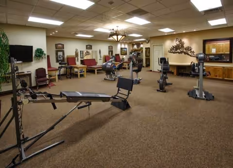 A spacious exercise room in a senior living facility with various fitness equipment including rowing machines, stationary bikes, and weight benches. The room has beige carpet, cream-colored walls, overhead fluorescent lighting, and framed artwork on the walls. There is a windowed office area on the right side of the room.