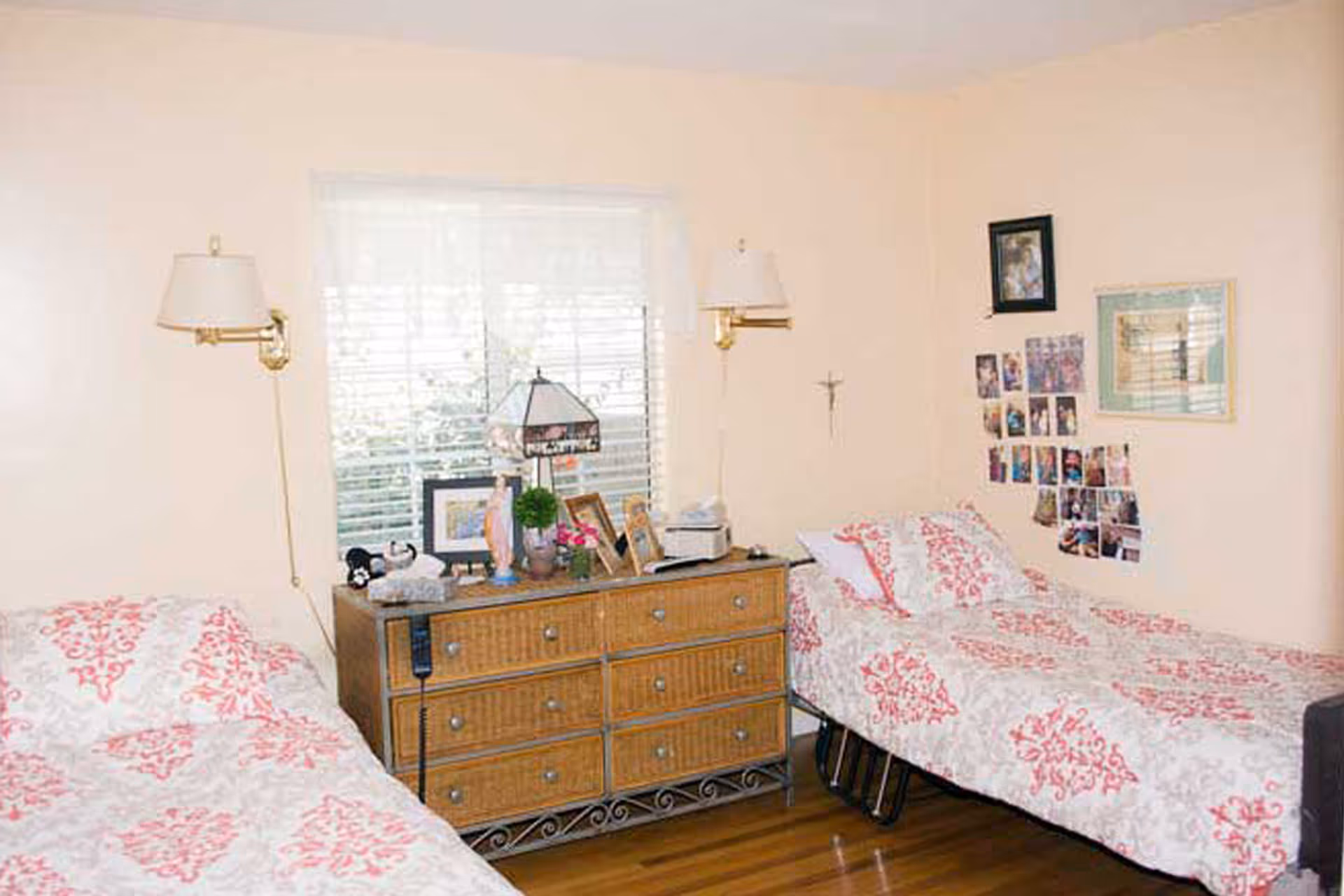 A bedroom with two twin beds featuring matching white and pink patterned bedspreads. Between the beds is a wooden dresser with six drawers, decorated with framed photos, a lamp, and small plants. The walls are light-colored and adorned with several photos and a crucifix. A window with white blinds is centered above the dresser.