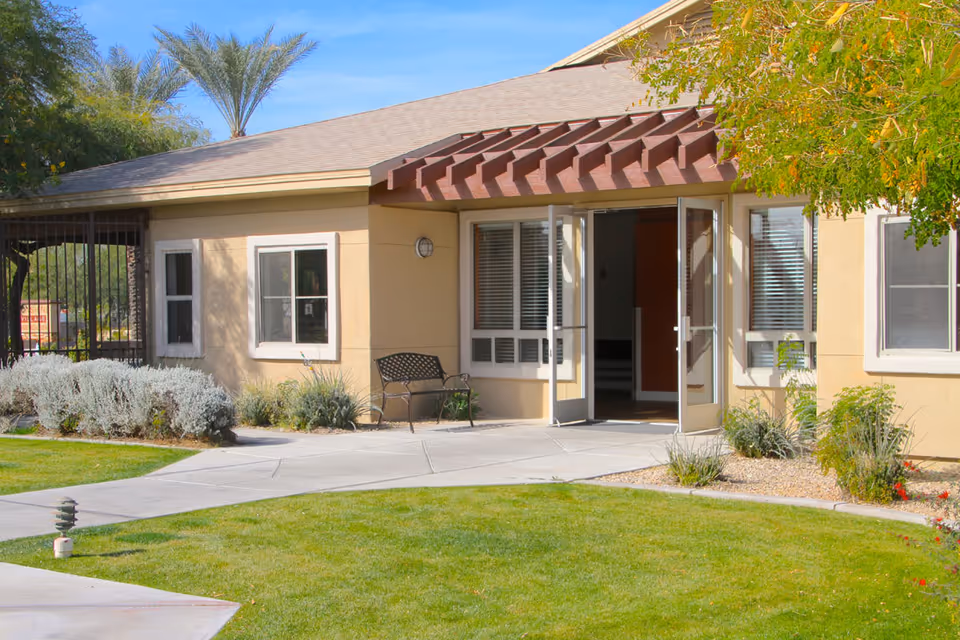 Exterior view of a single-story building with beige walls and a brown pergola over the entrance. The entrance has double glass doors that are open, and there are windows on either side. A metal bench is placed near the entrance, and there is a well-maintained lawn with shrubs and trees around the walkway leading to the door.