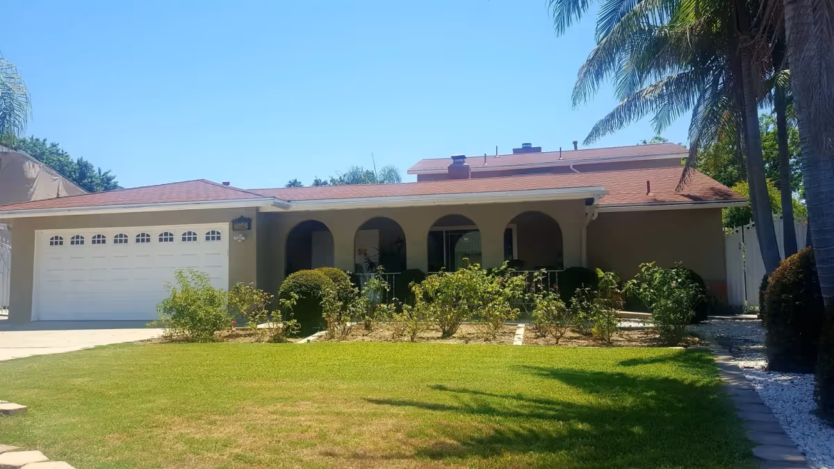 Exterior view of a single-story residential building with a red roof, beige walls, and a white garage door. The front yard features a well-maintained lawn, bushes, and palm trees under a clear blue sky.