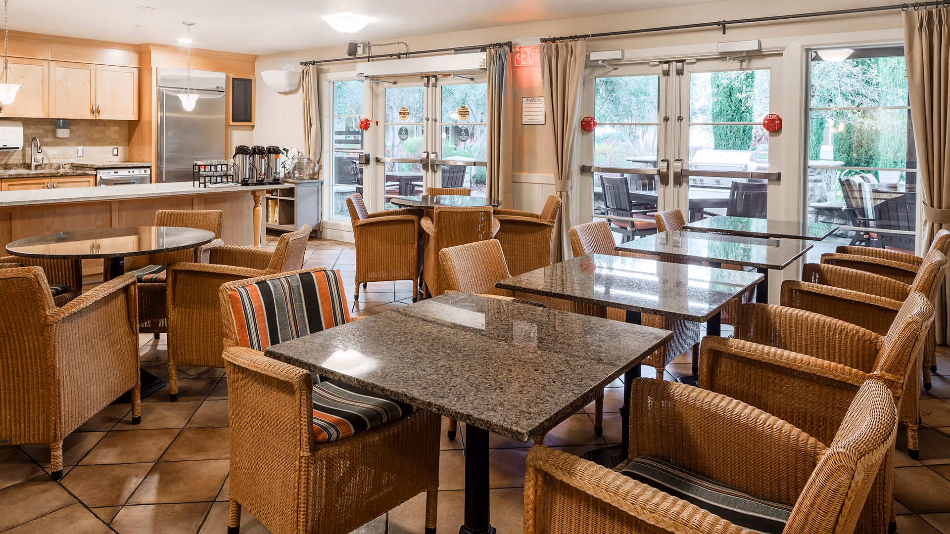 A bright dining area with several granite-top tables surrounded by wicker chairs with striped cushions. The room has large glass doors leading to an outdoor patio with more seating. The kitchen area with wooden cabinets and stainless steel appliances is visible in the background.