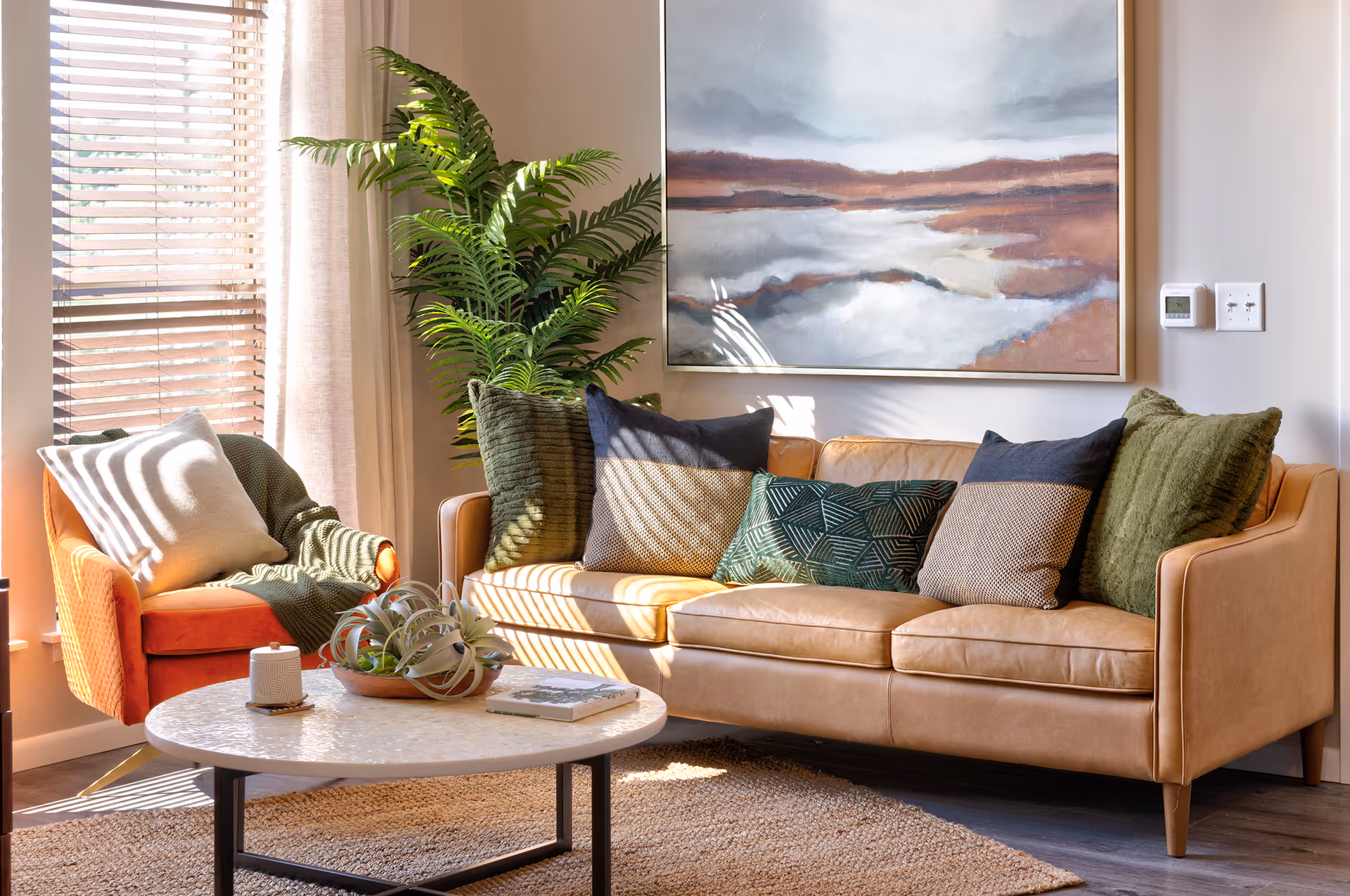 A cozy living room with a tan leather sofa adorned with multiple green and patterned throw pillows. To the left, there is an orange armchair with a white pillow and a green throw blanket. A round white coffee table with a small plant and a book sits in front of the sofa. Behind the sofa, a large framed landscape painting hangs on the wall, and a tall green plant is placed near the window with blinds and light curtains allowing sunlight to stream in.