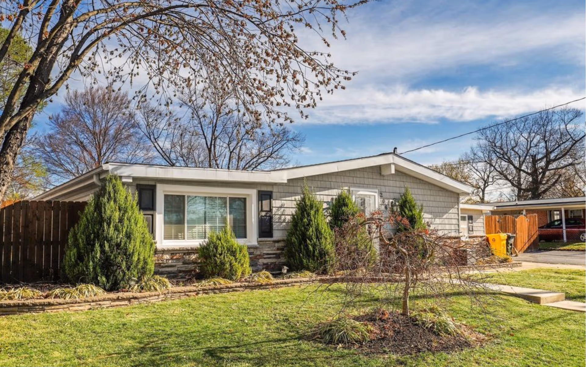 Single-story residential building with gray siding and a white-framed window, surrounded by green shrubs and a small tree in the front yard under a partly cloudy blue sky.