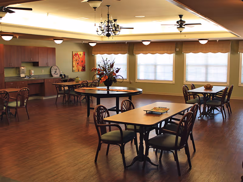 A spacious dining room with several wooden tables and chairs arranged neatly. The room features large windows with blinds, a chandelier, ceiling fans, and a cabinet area with a countertop along one wall. A floral centerpiece is placed on a round table in the middle of the room.