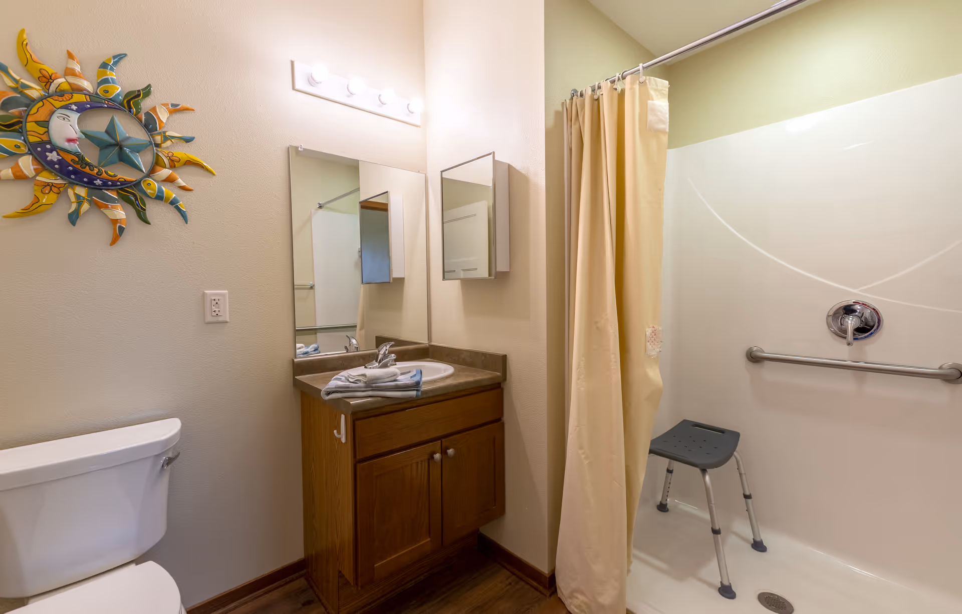 Accessible bathroom with a vanity and mirror, toilet, and a shower with a curtain, grab bar, and shower stool.