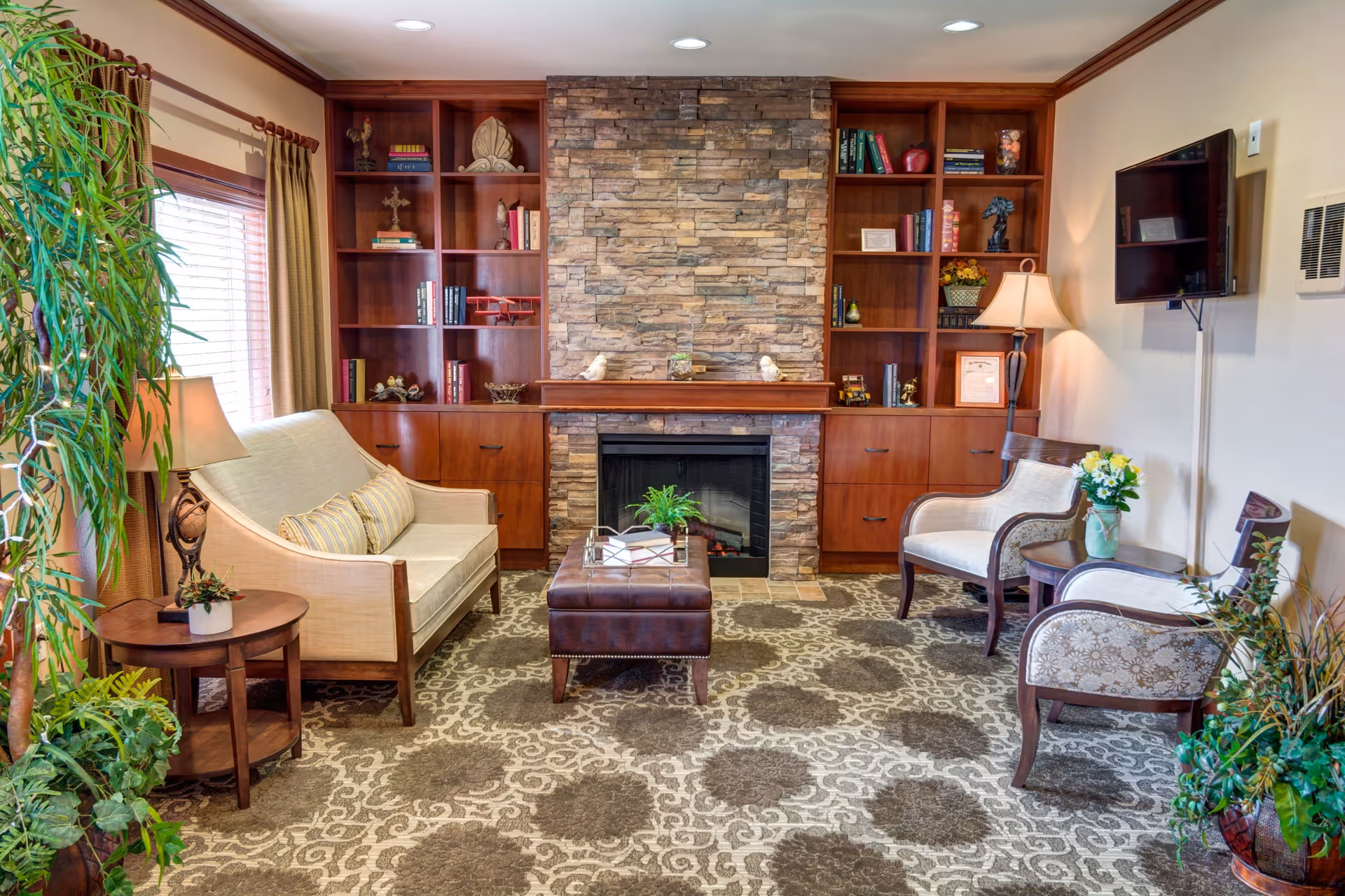 A cozy living room with a stone fireplace centered between two wooden built-in bookshelves filled with books and decorative items. The room features a beige loveseat with striped pillows, two upholstered armchairs, a brown leather ottoman, a side table with a lamp, and several potted plants. A flat-screen TV is mounted on the wall to the right.