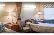 A cozy bedroom in a senior living facility featuring a single bed with beige bedding and decorative pillows, a wooden nightstand with a lamp, a chair, and a window with a patterned valance and closed blinds.