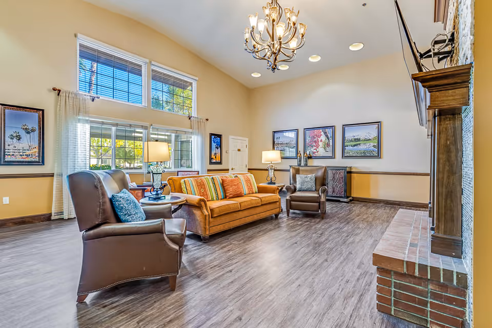 A bright and spacious living room with high ceilings, featuring a brown leather armchair, an orange sofa with striped cushions, a side table with a lamp, and a brick fireplace with a mounted TV above it. Large windows with white curtains allow natural light to fill the room, and framed artwork decorates the walls.