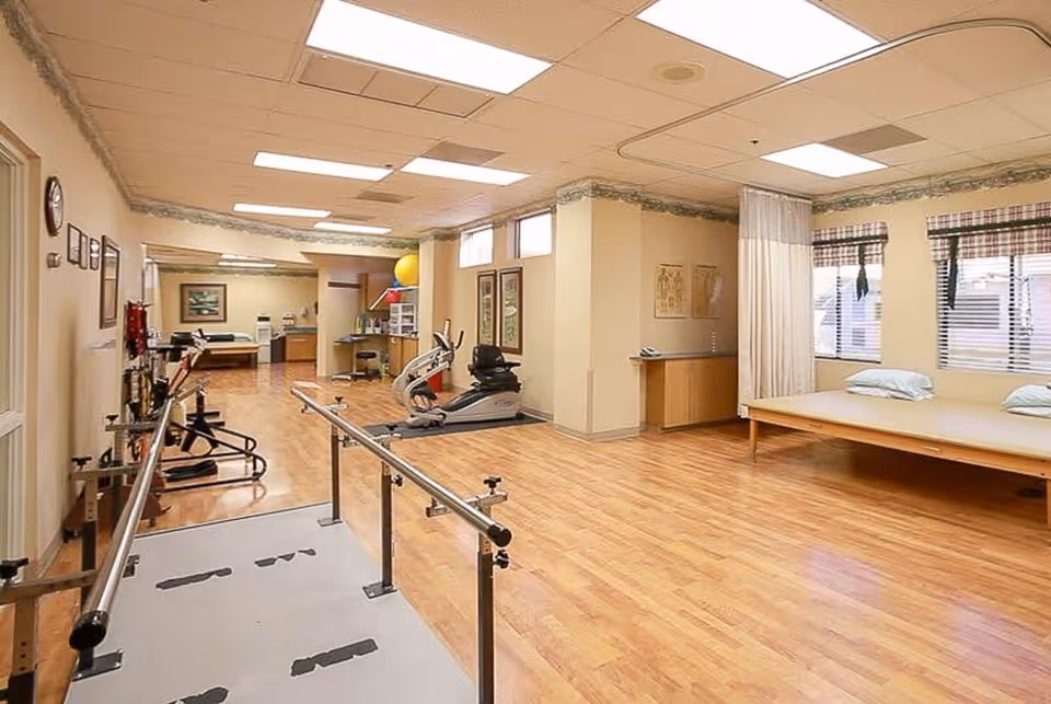 A spacious rehabilitation room with wooden flooring featuring parallel bars for walking exercises, an exercise bike, a therapy bed with pillows, and various medical and therapy equipment. The room has large windows with blinds and plaid valances, fluorescent ceiling lights, and light-colored walls with decorative border trim.