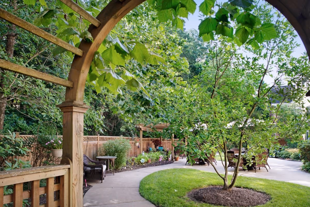 A peaceful outdoor garden area at Sunrise of Newtown Square featuring a wooden archway, a curved concrete pathway, green grass, various trees and plants, and outdoor seating with wicker chairs and tables.