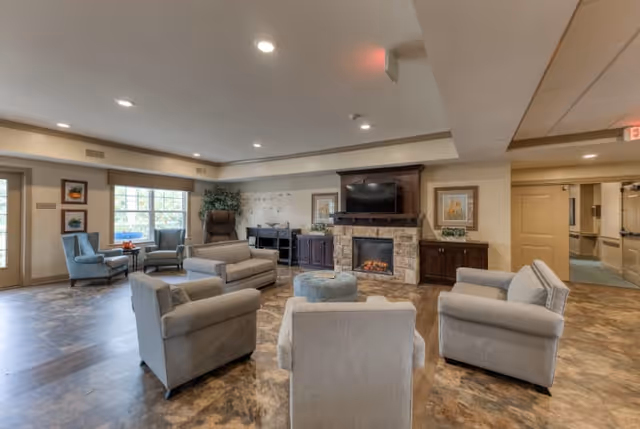 A spacious senior living facility common area with several comfortable armchairs arranged around a central ottoman. The room features a stone fireplace with a mounted flat-screen TV above it, framed artwork on the walls, large windows letting in natural light, and a door leading outside. The flooring is a wood-like pattern, and the ceiling has recessed lighting.