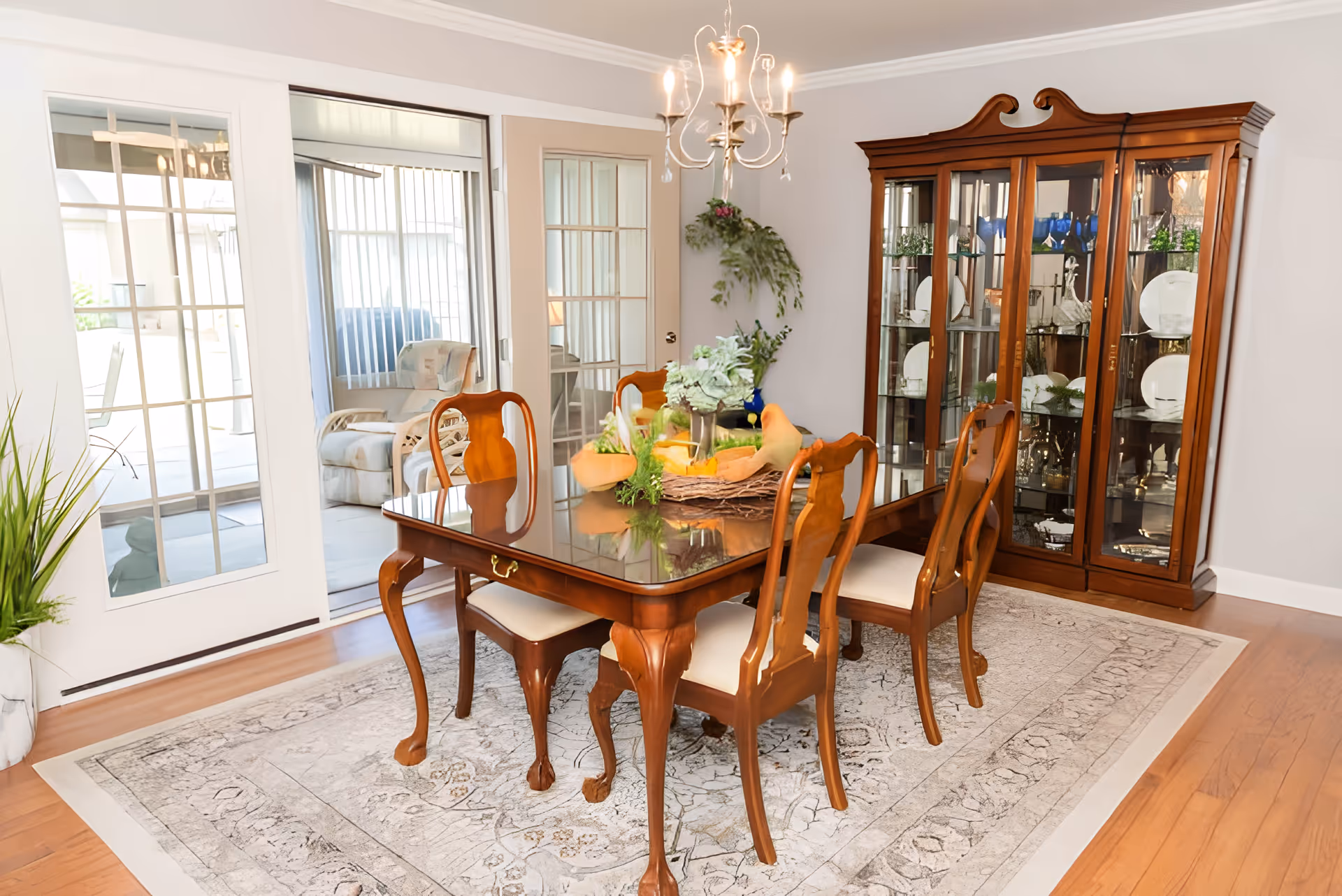 A traditional dining room with a polished wooden dining table and six matching chairs with white cushions. The table is decorated with a centerpiece featuring greenery and decorative birds. Behind the table is a large wooden china cabinet displaying plates and glassware. The room has hardwood floors with a patterned area rug underneath the table. French doors and sliding glass doors lead to an adjacent sunroom with a cushioned armchair visible. A chandelier with lit bulbs hangs above the dining table.