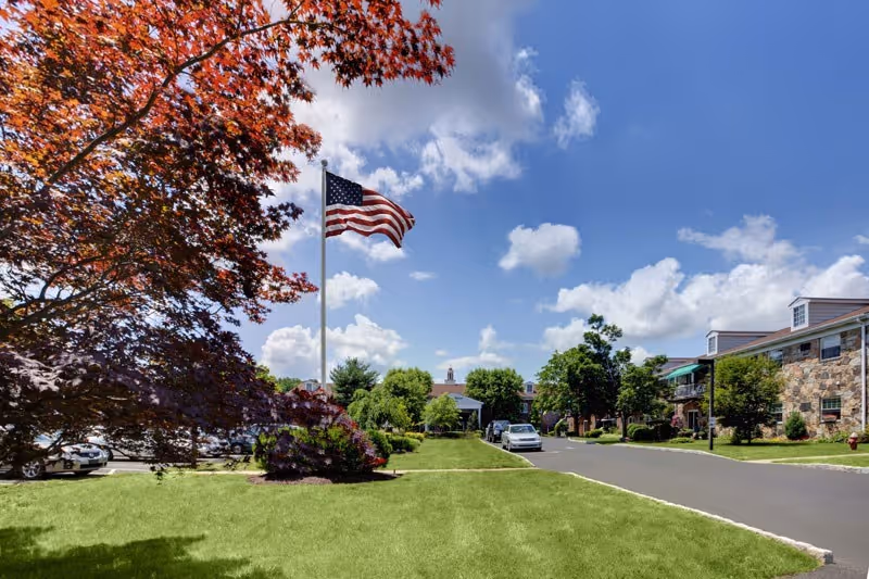 American flag on a pole in front of a senior living community with a manicured lawn, driveway, trees, and buildings under a blue sky.