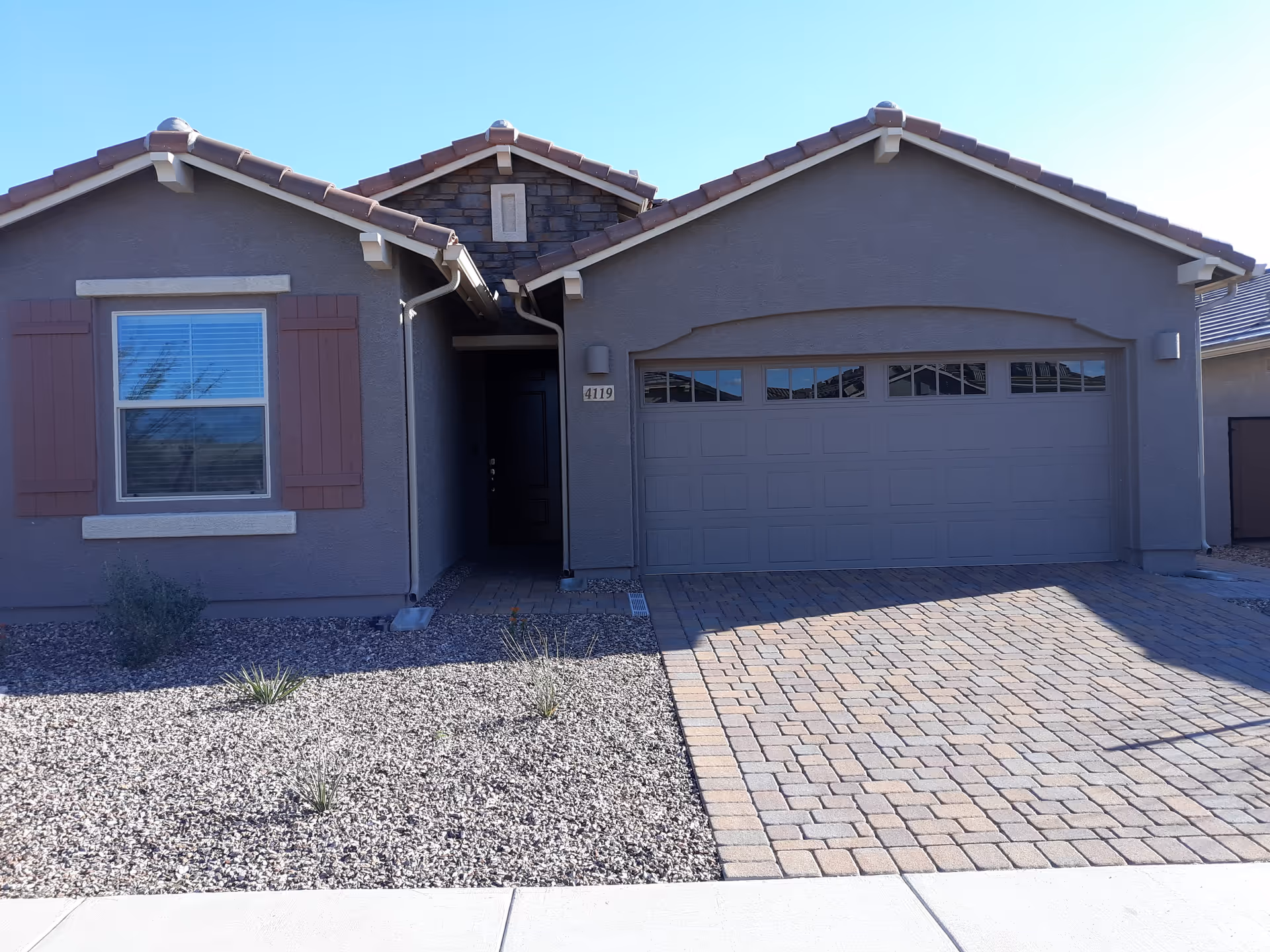 Front exterior view of a single-story house with a two-car garage, a window with shutters, and a gravel yard with sparse desert plants under a clear blue sky.