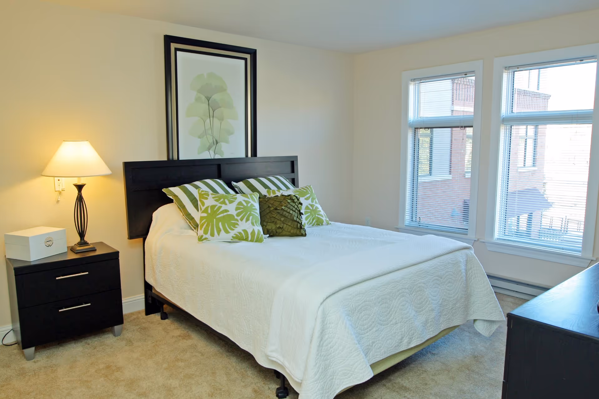 A bright bedroom with a neatly made bed featuring white bedding and green decorative pillows. There is a black headboard and a framed botanical print above the bed. To the left of the bed is a black nightstand with a lamp and a white box. Two large windows with blinds allow natural light into the room.