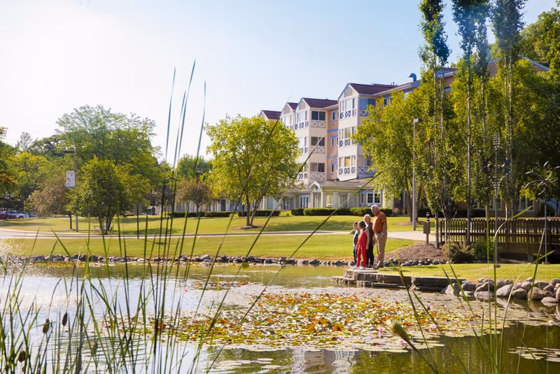A serene outdoor scene at Meth-Wick Community featuring a pond with lily pads in the foreground, a family of four standing on a stone platform by the water, and a multi-story residential building surrounded by green trees and a well-maintained lawn in the background under a clear sky.