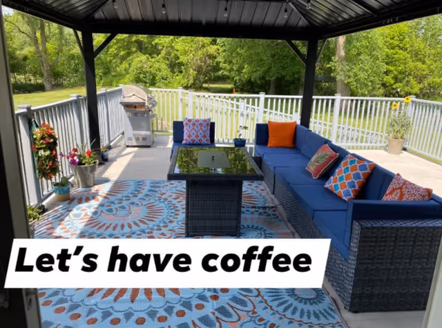 Covered outdoor patio with a blue wicker sectional sofa, colorful pillows, a glass-top table, patterned rug, potted plants and a grill overlooking a grassy yard.