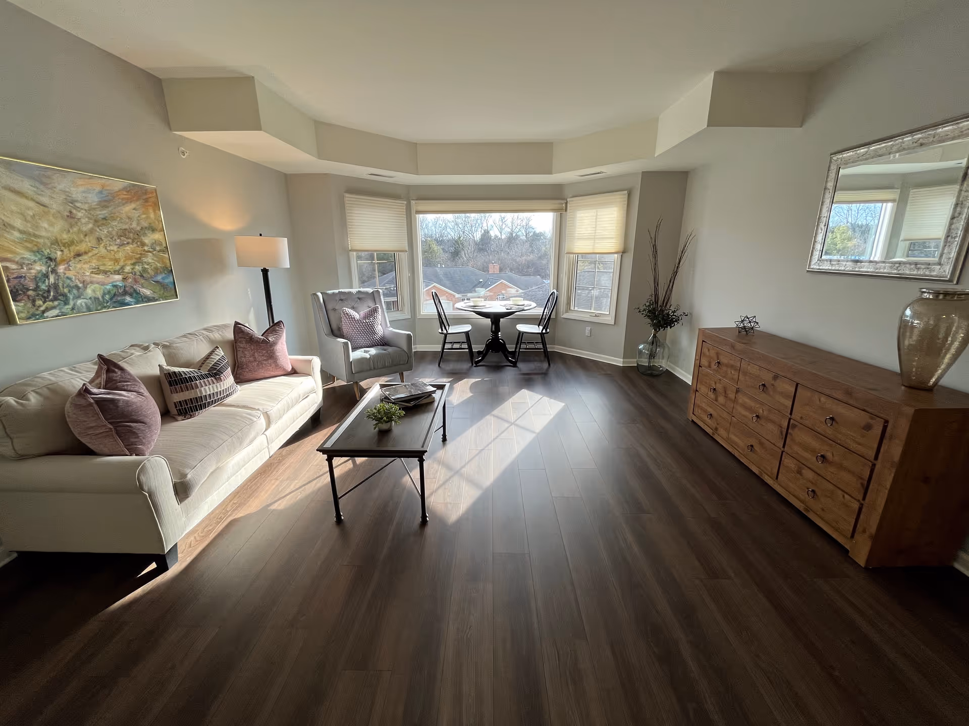 A bright living room with a beige sofa adorned with purple and patterned pillows, a gray armchair, a wooden coffee table with a small plant, and a round dining table with two chairs near a large bay window. The room has wooden flooring, a wooden dresser with decorative items, a floor lamp, a large mirror on the wall, and a colorful painting.