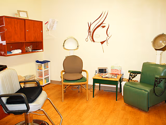 Interior of a small salon or beauty room with three chairs including a salon chair, a brown cushioned chair, and a green vintage salon chair with a hair dryer hood. There is a small table with magazines and tissue box, a wall decal of a woman's face with flowing hair, wooden cabinets on the wall, and a plastic drawer organizer in the corner.