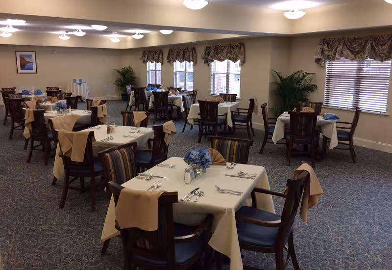A dining room in a senior living facility with multiple tables covered in white tablecloths and beige napkins draped over the chairs. Each table is set with silverware, salt and pepper shakers, and a centerpiece of blue flowers. The room has carpeted floors, several windows with blinds and valances, and potted plants near the walls.