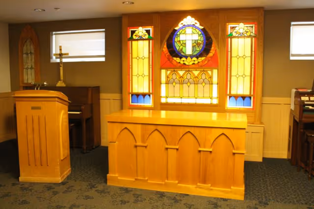Interior of a chapel or worship room with a wooden podium, an altar, a piano, and a stained glass window featuring a cross and decorative patterns. The room has carpeted flooring and beige walls with two small rectangular windows.