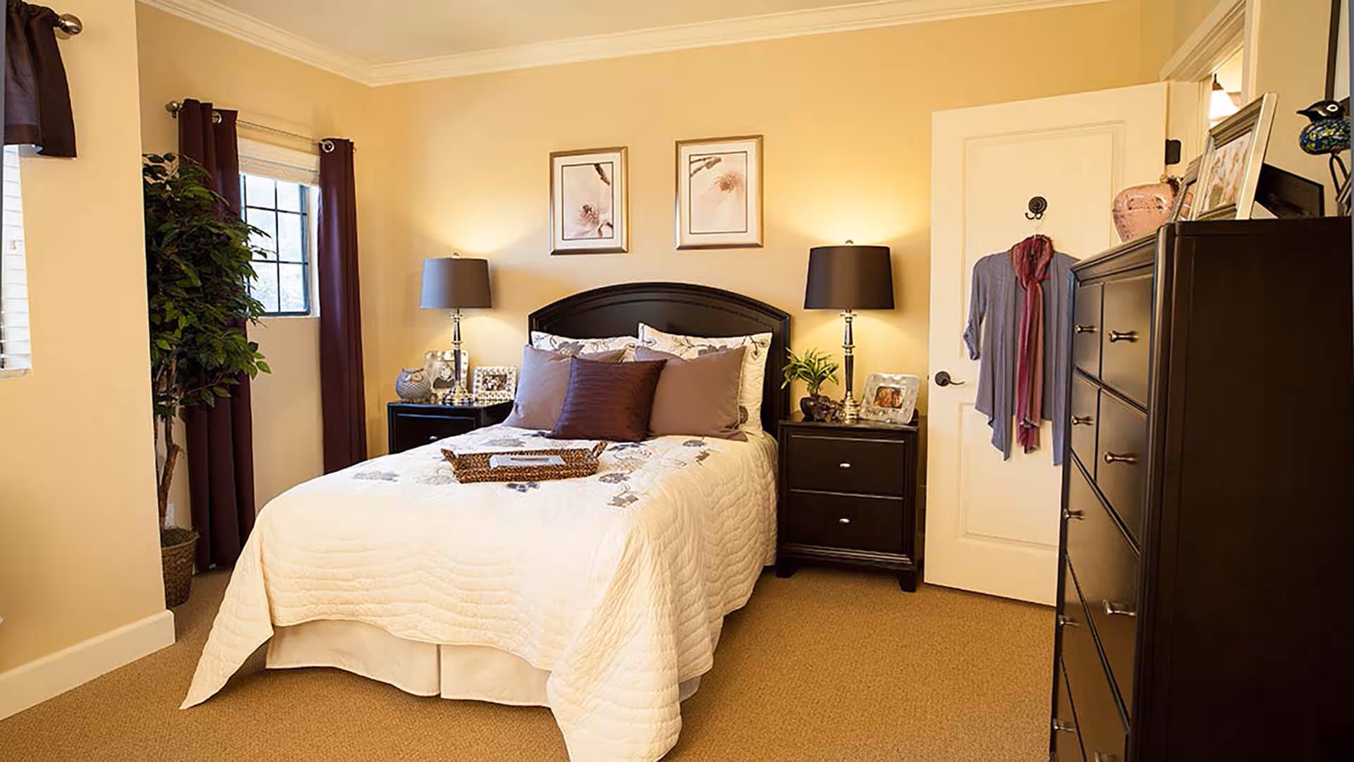 A cozy bedroom with a neatly made bed featuring a white quilt and several pillows. There are two dark bedside tables with matching lamps, picture frames, and decorative items. A window with dark curtains and a potted plant are on the left side. A dresser with framed photos and a decorative bird is on the right, and a door with clothes hanging on a hook is visible in the background.
