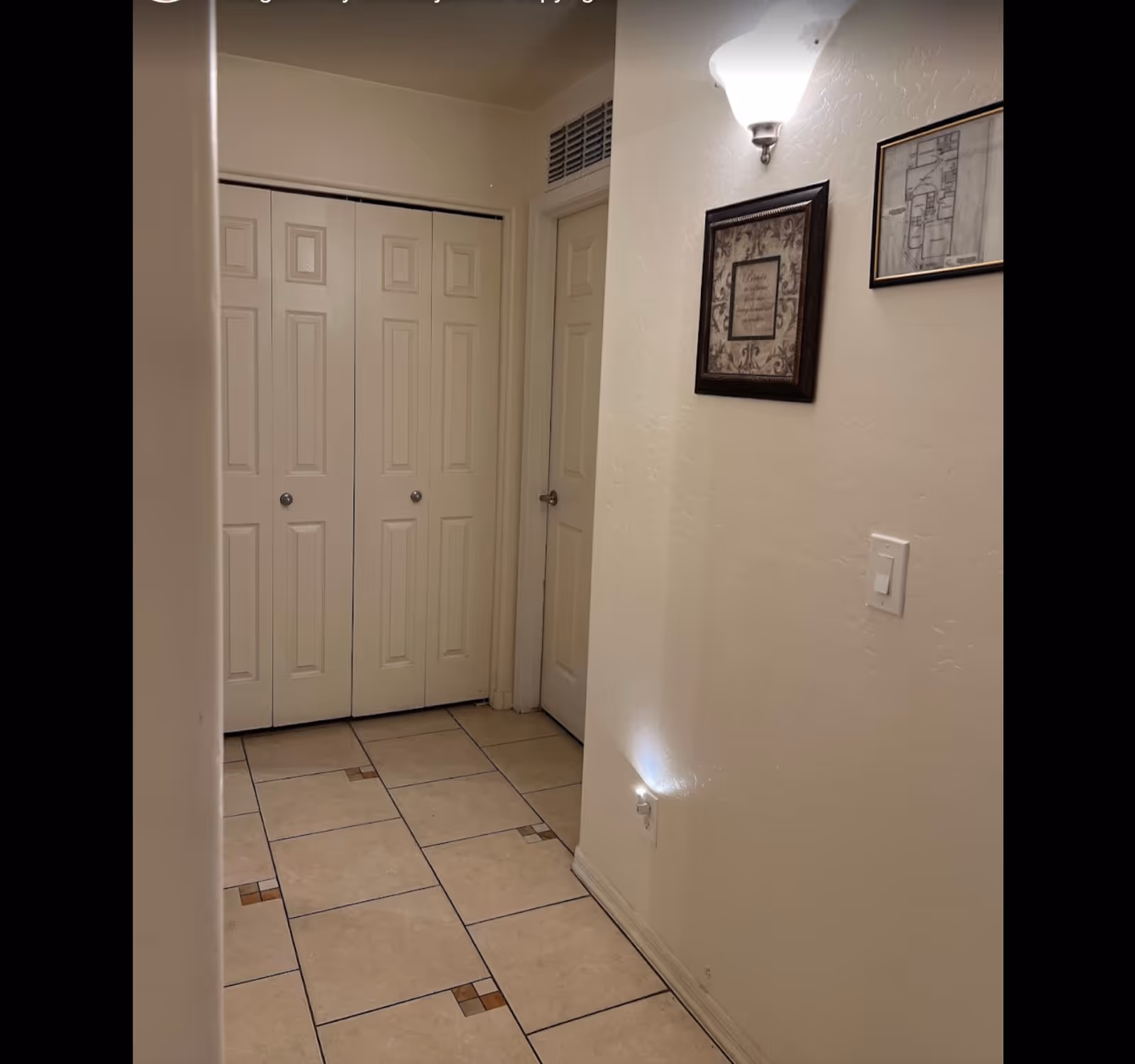 A hallway with beige tiled floor featuring small square accent tiles, cream-colored walls, and two closed doors. One door is a double closet door with panels, and the other is a single door with a silver handle. On the right wall, there is a light switch, a wall-mounted light fixture, and two framed pictures, one of which appears to be a floor plan.
