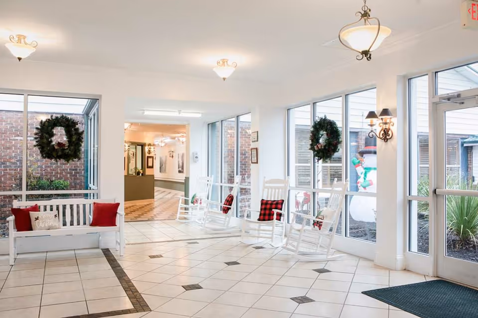 Bright lobby seating area with white rocking chairs, a white bench with red pillows, and holiday wreaths on large windows.