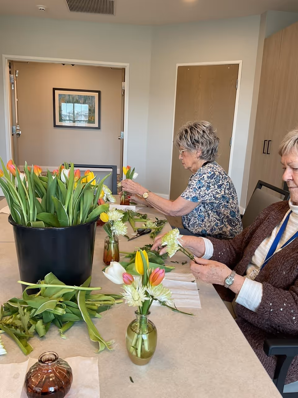 Two elderly women sitting at a table arranging flowers in small vases. The table has a large black container filled with tulips and various flower stems and leaves scattered around. The room has light-colored walls, a wooden door, and a framed picture on the wall.