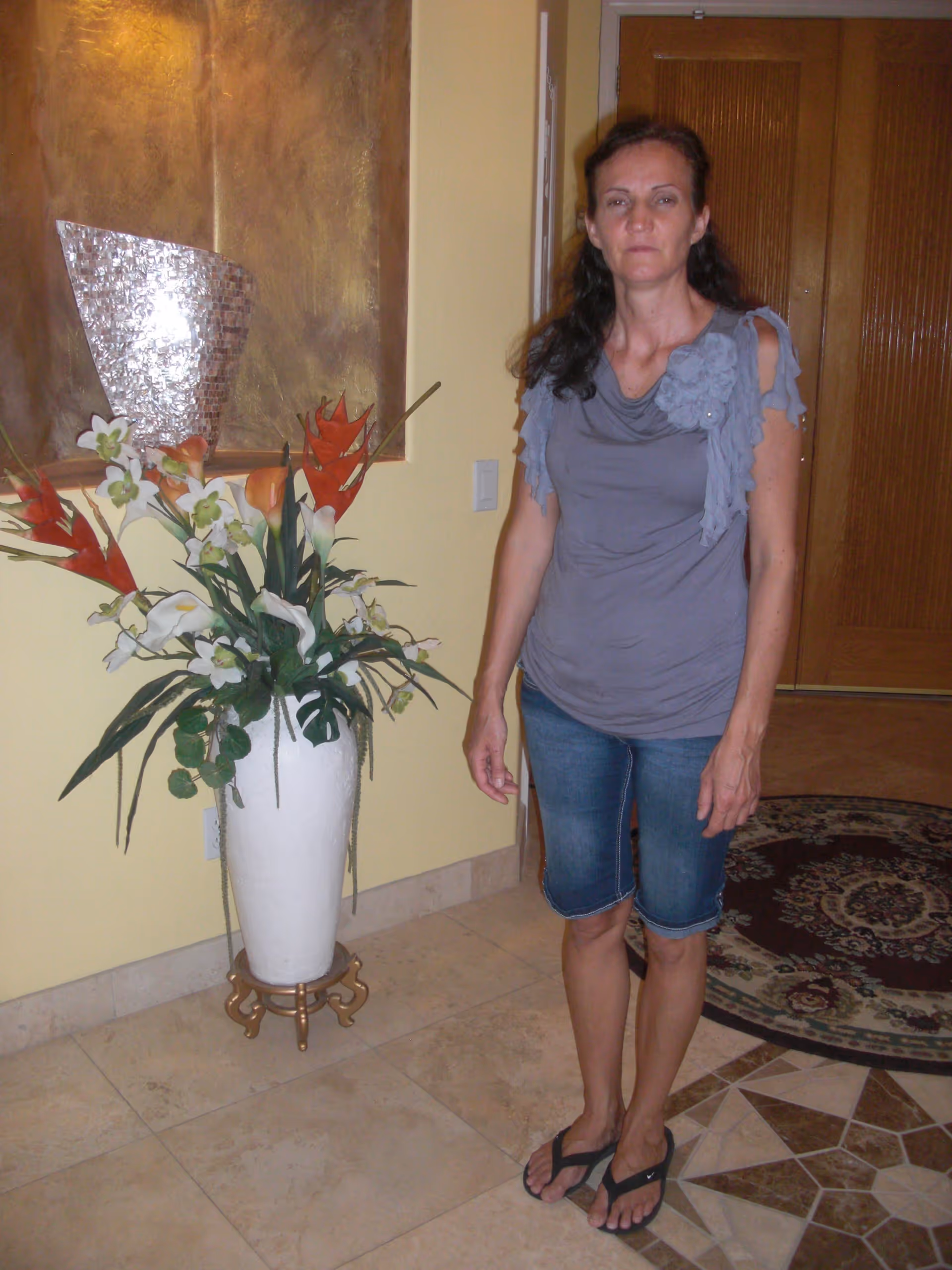 A woman standing indoors near a large white vase with an arrangement of artificial flowers. The background includes a yellow wall, a decorative wall panel, a wooden door, and a patterned rug on the tiled floor.
