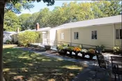 Single-story light-colored building with a wheelchair ramp, potted flowers and outdoor seating on a shaded lawn.