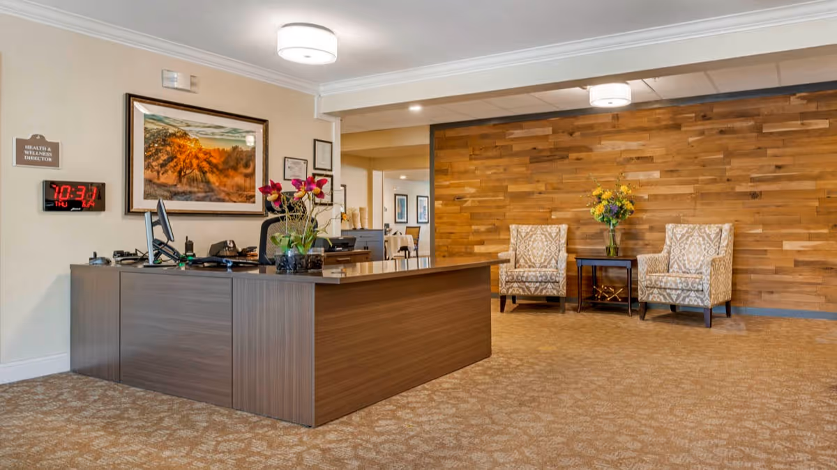 Reception area with a wooden desk, two computer monitors, a vase with flowers, and office supplies. Behind the desk is a wall with a framed landscape picture and a digital clock displaying the time 10:31. To the right, there are two patterned armchairs with a small table between them holding a vase of flowers, set against a wooden accent wall.