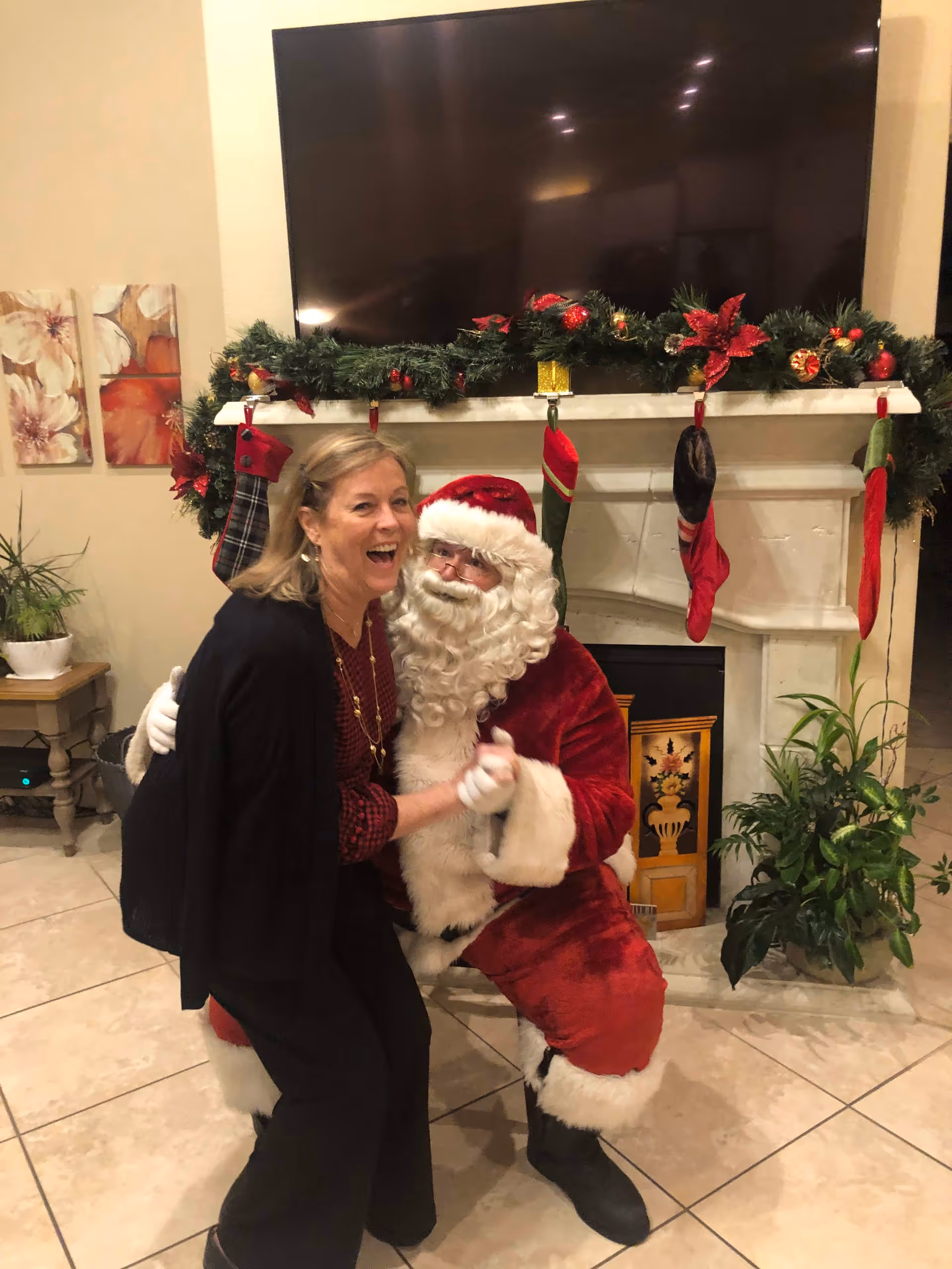A woman laughing and posing with a person dressed as Santa Claus in front of a decorated fireplace with Christmas stockings and garland. There is a large TV mounted above the fireplace and some plants and artwork in the background.
