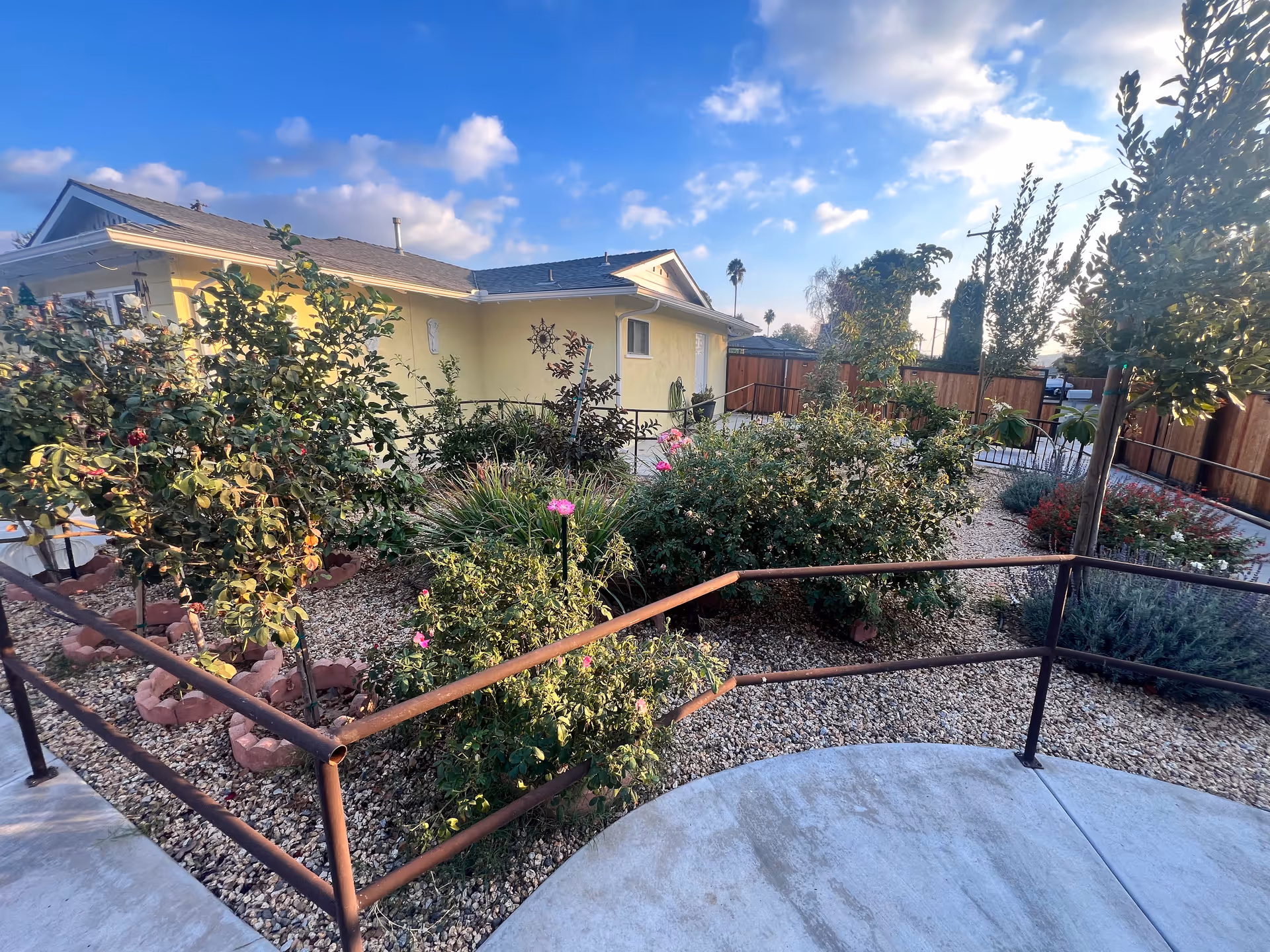 Front yard and entrance ramp of a single-story yellow residence with landscaped gravel beds, shrubs, and a metal handrail.