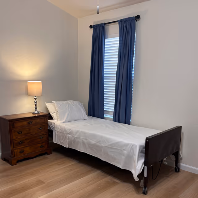A simple bedroom with a single bed covered in white sheets and pillows, a wooden nightstand with a lit lamp, and a window with blue curtains and white blinds.