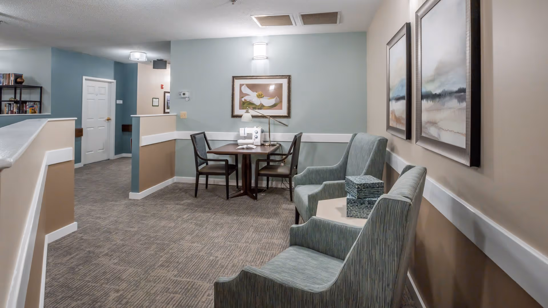 A cozy interior corner of a senior living facility with two upholstered armchairs and a small table between them holding decorative boxes. On the opposite wall, there is a small round table with two chairs and a sewing machine on top. The walls are painted in soft beige and light blue tones, decorated with framed artwork. The carpeted floor and soft lighting create a warm and inviting atmosphere.