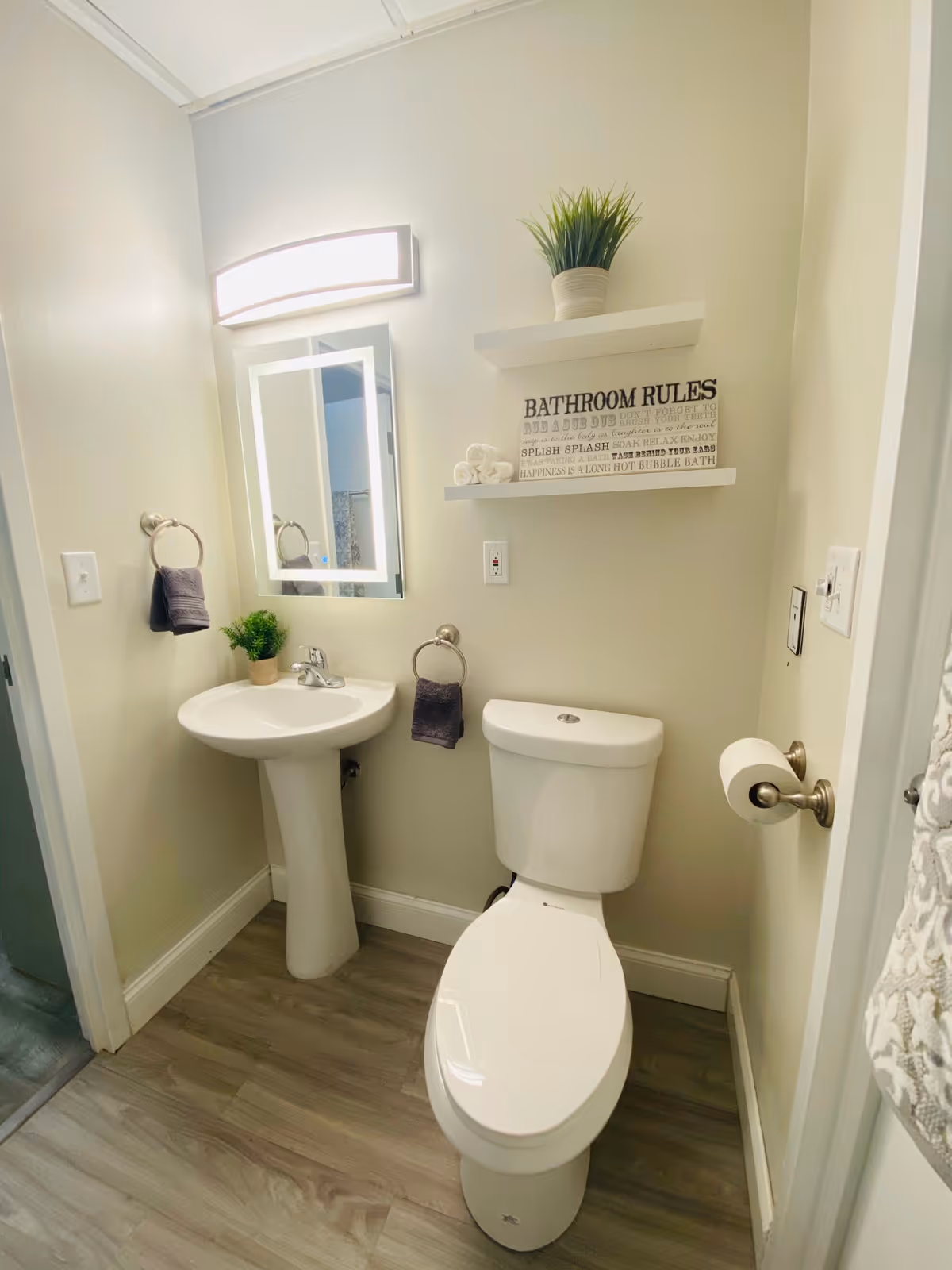 A clean and modern bathroom featuring a white pedestal sink with a small green plant on it, a white toilet, two towel rings each holding a dark gray hand towel, and two white floating shelves on the wall. The top shelf has a potted green plant, and the lower shelf has rolled white towels and a decorative sign that reads 'BATHROOM RULES'. The floor is wood-style laminate, and the walls are painted light beige. A rectangular mirror with built-in lighting is mounted above the sink.