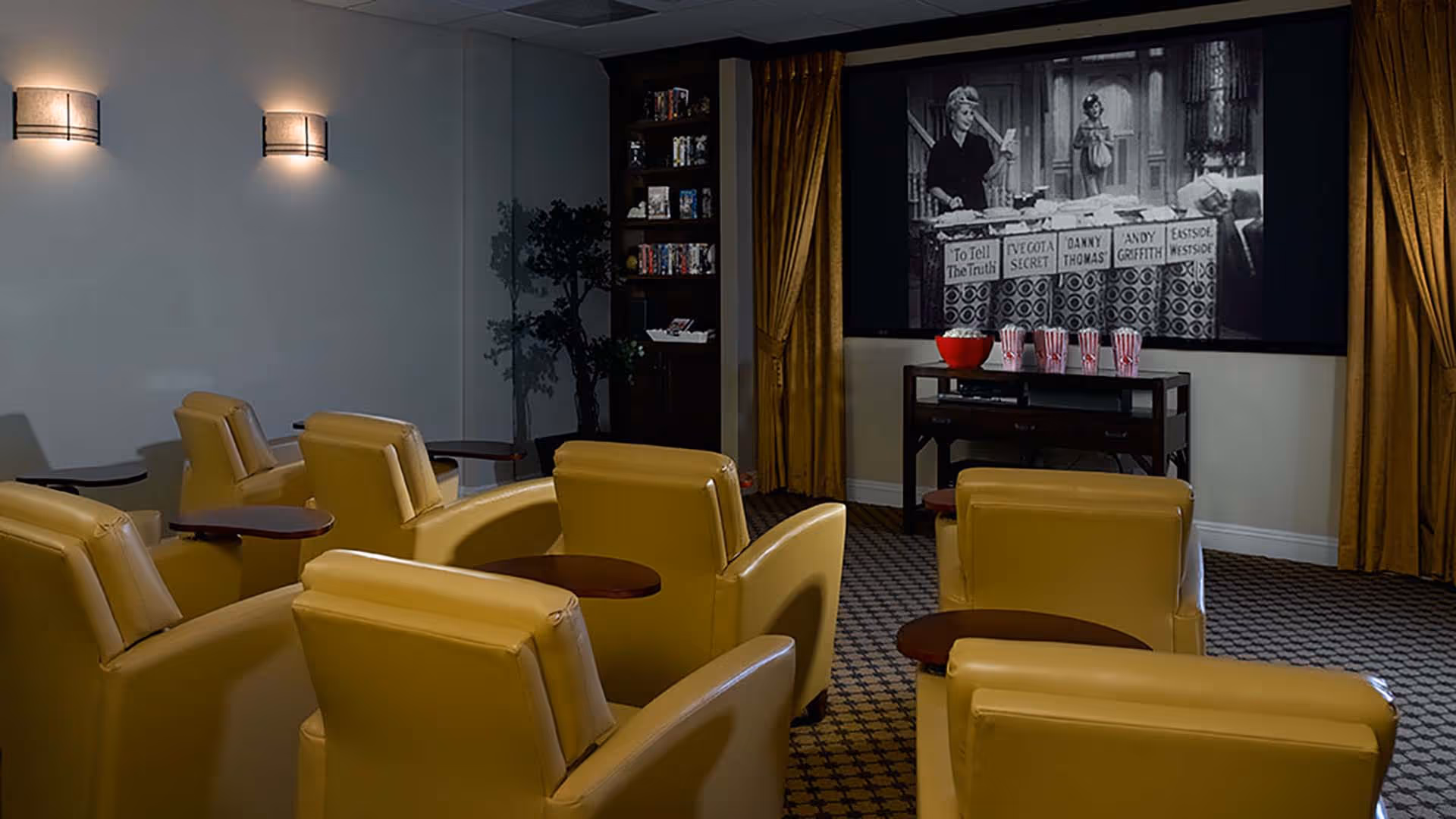 A cozy movie theater room with several yellow armchairs arranged in rows facing a large screen showing a black and white film. There are small wooden tables attached to some chairs, a popcorn bowl and striped popcorn containers on a table beneath the screen, and warm lighting on the walls. The room has patterned carpet and gold curtains framing the screen.