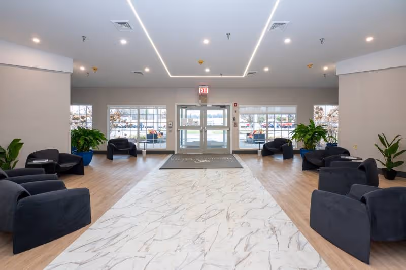 A spacious and well-lit lobby area with a marble-patterned floor runner leading to glass double doors. The room features several black cushioned chairs arranged in pairs along the sides, with green potted plants placed near the walls. Large windows on either side of the doors allow natural light to enter, and the ceiling has modern recessed lighting with a geometric design.