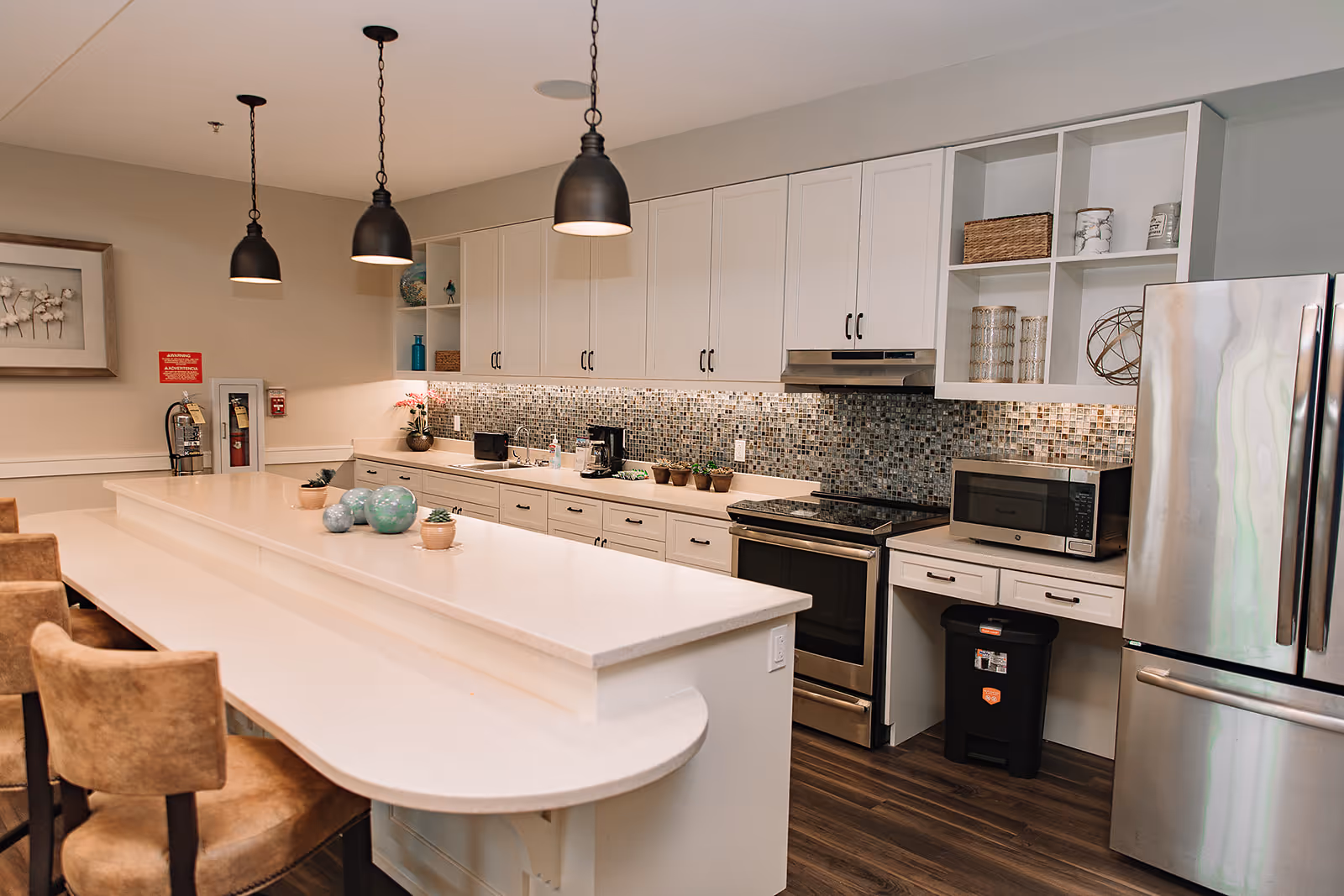 A modern kitchen area with a long white countertop island surrounded by brown cushioned chairs. The kitchen features white cabinets, a mosaic tile backsplash, stainless steel appliances including a refrigerator and oven, a microwave, and various decorative items on the counter and shelves. Three black pendant lights hang from the ceiling above the island.