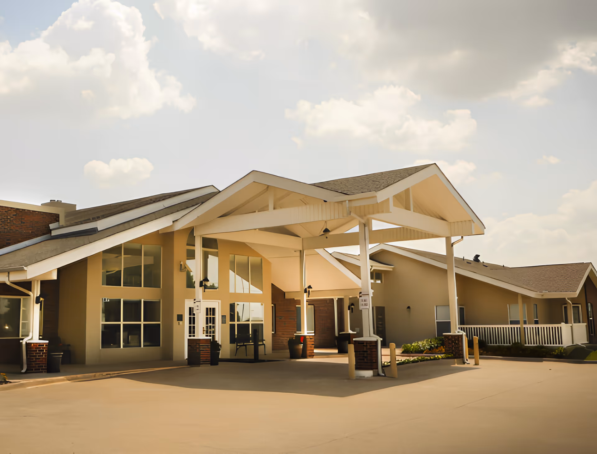 Exterior view of a single-story building with a covered entrance supported by white pillars. The building has large windows and a sloped roof under a partly cloudy sky.