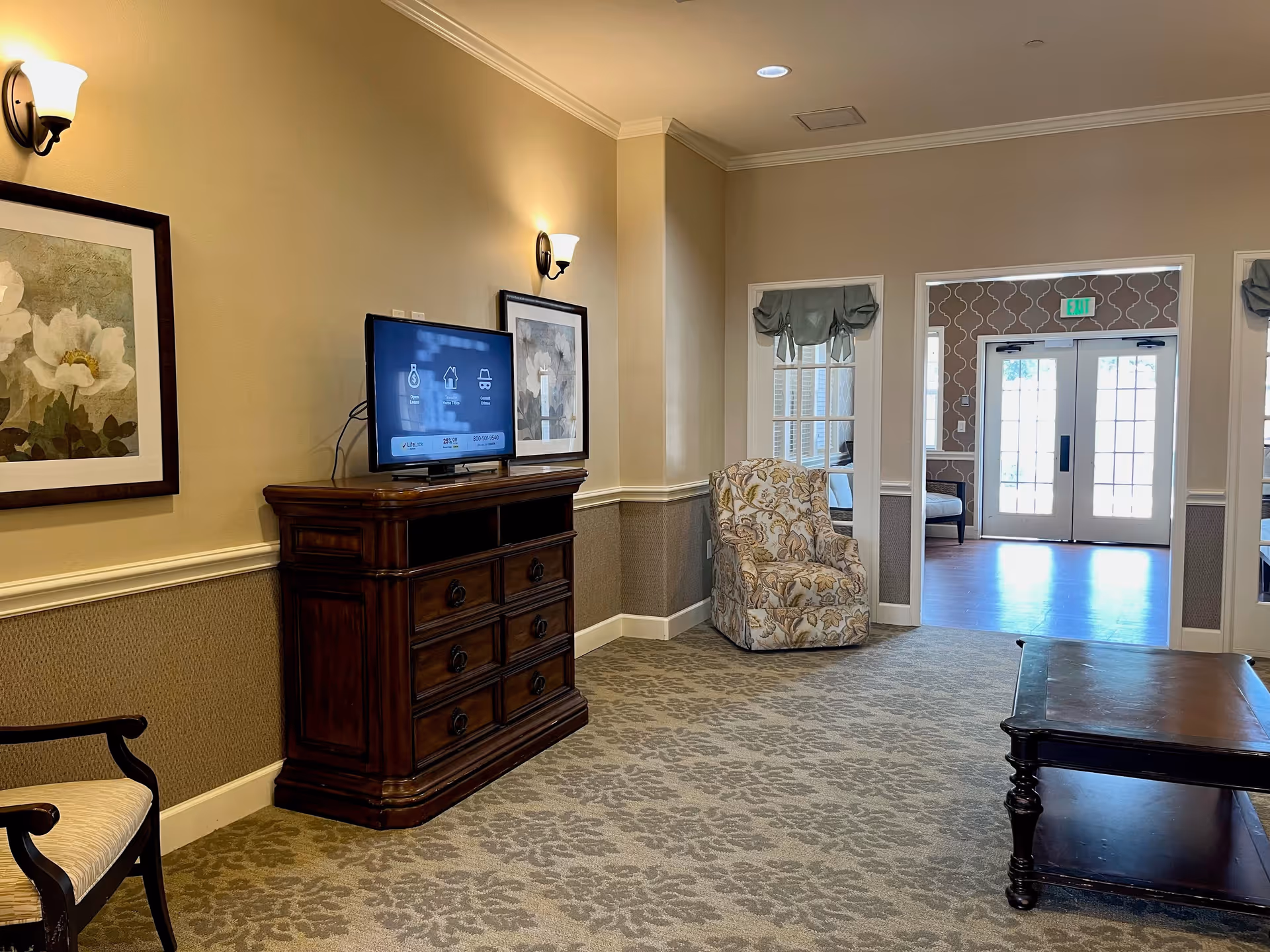A cozy living room area in a senior living facility with beige walls and patterned carpet. The room features a wooden dresser with a flat-screen TV on top, two framed floral paintings on the wall, a floral upholstered armchair, a wooden coffee table, and an armchair with light upholstery. There are two windows with green valances and a double glass door leading to another room with patterned wallpaper and additional seating.