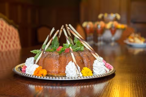 A close-up of a decorated bundt cake on a silver platter, garnished with whipped cream, colorful flowers, and chocolate sticks, placed on a polished wooden table in a warmly lit room.