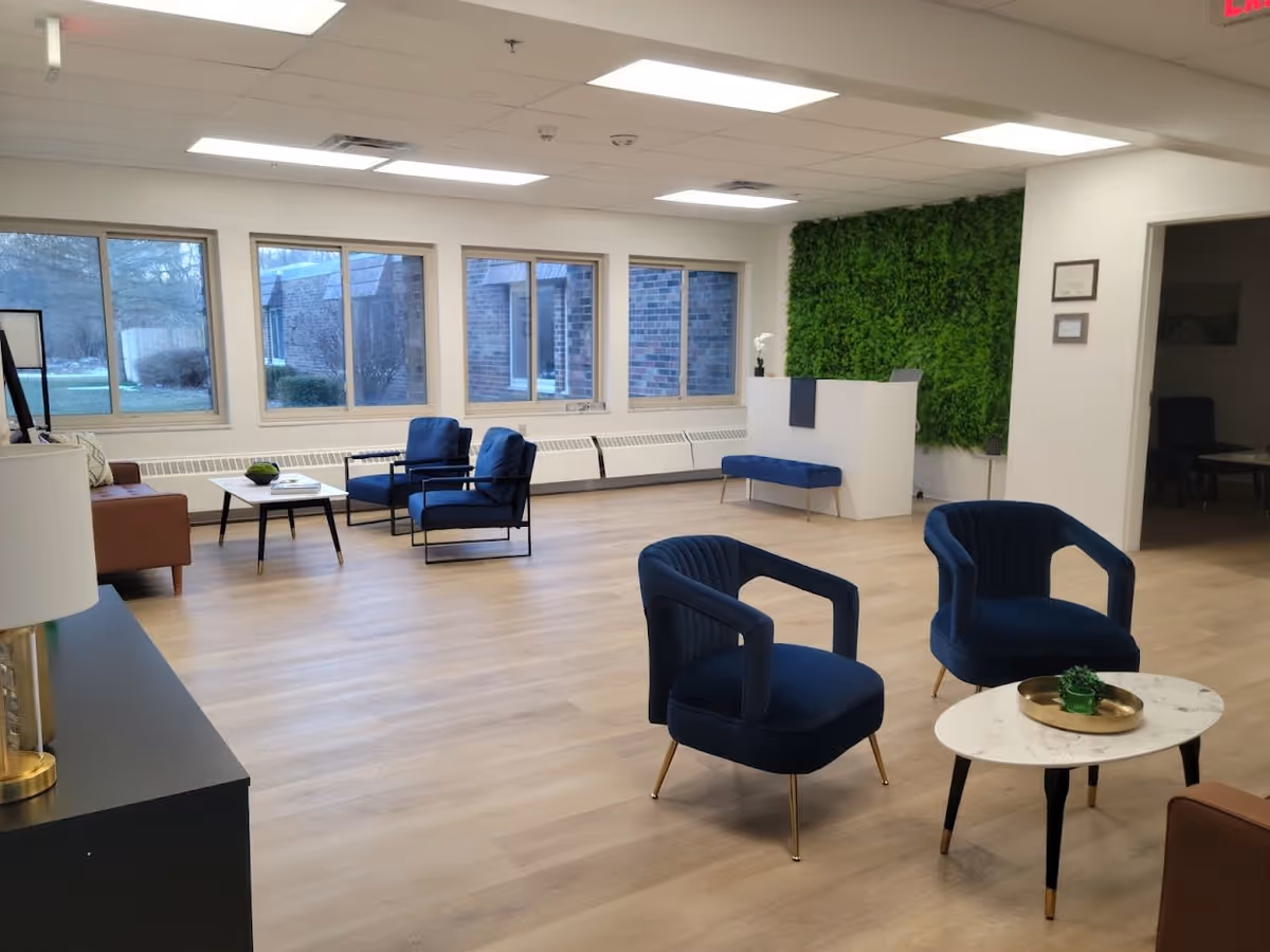 A spacious, modern living room area with light wood flooring, large windows letting in natural light, and a green plant wall behind a white reception desk. The room features blue upholstered chairs, a small white marble table with a decorative tray and plant, and a brown sofa with a coffee table. The ceiling has recessed lighting and there is an exit sign visible in the top right corner.