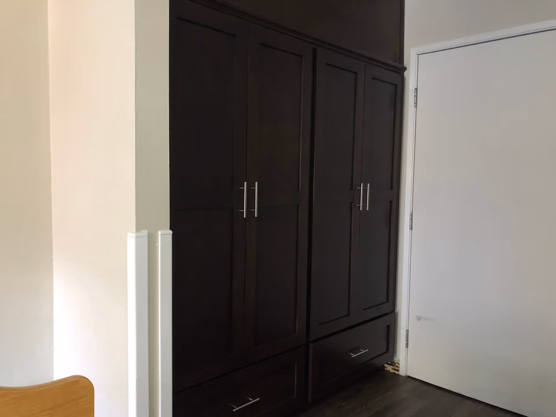 A corner of a room featuring a dark wooden cabinet with multiple doors and drawers, next to a white door with metal hinges. The floor is dark wood, and part of a light-colored wall and a wooden chair are visible on the left side.