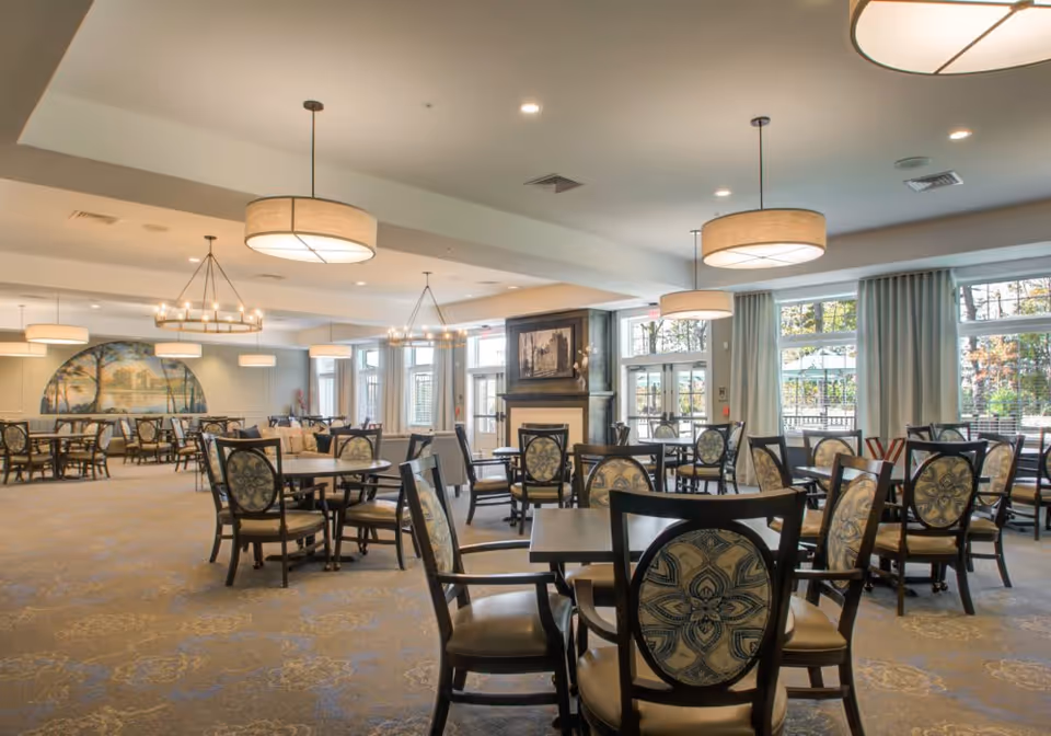 A spacious dining room with multiple round tables and chairs featuring patterned upholstery. The room is well-lit with large windows allowing natural light and several hanging light fixtures. There is a fireplace with a framed picture above it and a large mural on one wall.