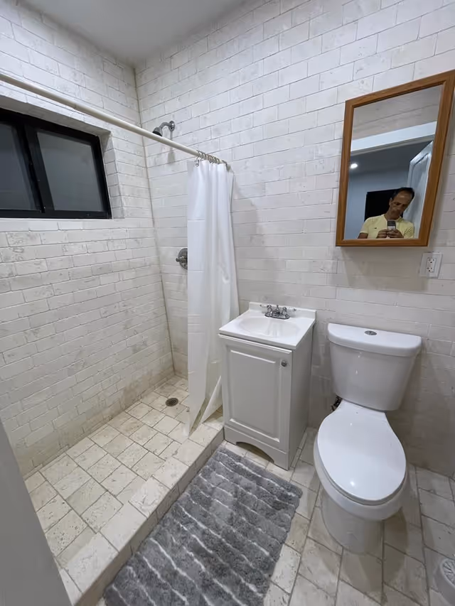Small white-tiled bathroom with a shower and curtain, a vanity sink, toilet, wall mirror reflecting a person, and a gray bath mat.