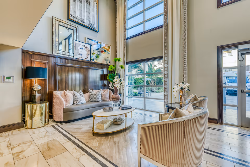 A bright and elegant senior living facility lounge area with a curved sofa adorned with multiple pillows, two armchairs, a round coffee table with decorative items, a tall floor lamp, and large windows letting in natural light. The walls are decorated with framed artwork and mirrors, and there is a view of an outdoor garden area through the windows.